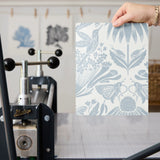 Symmetrical botanical pattern with stylized flowers, leaves, and moths in soft blue-gray on cream ba