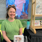 Woman in a green shirt holding a book, with art and a water bottle in the background
